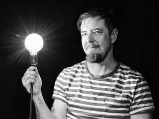 Adrien wears a stripped t-shirt and holds up a lit exposed lightbulb while looking at the camera